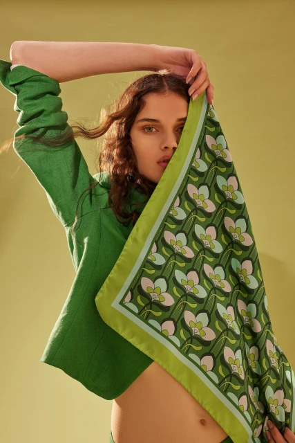 Brunette woman in a green outfit holds a floral-patterned scarf over her face, set against a green background in a fashion studio setting.