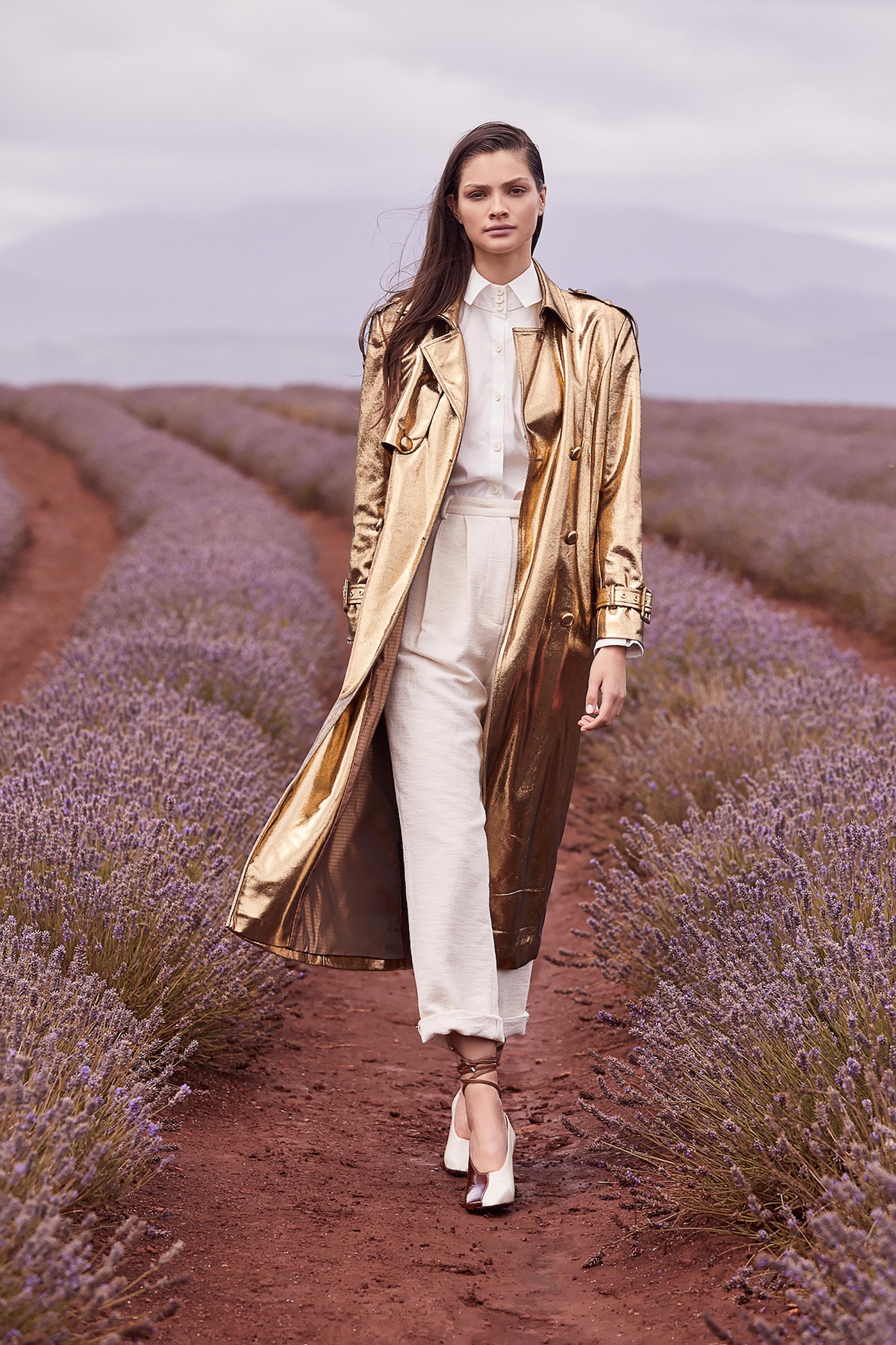 Woman in a gold trench coat walks through a lavender field. She wears white pants, a button-down shirt, and white high-heeled shoes.