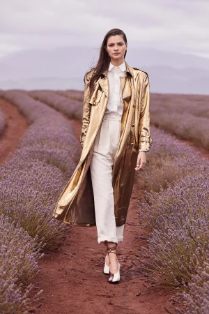 Woman in a gold trench coat walks through a lavender field. She wears white pants, a button-down shirt, and white high-heeled shoes.