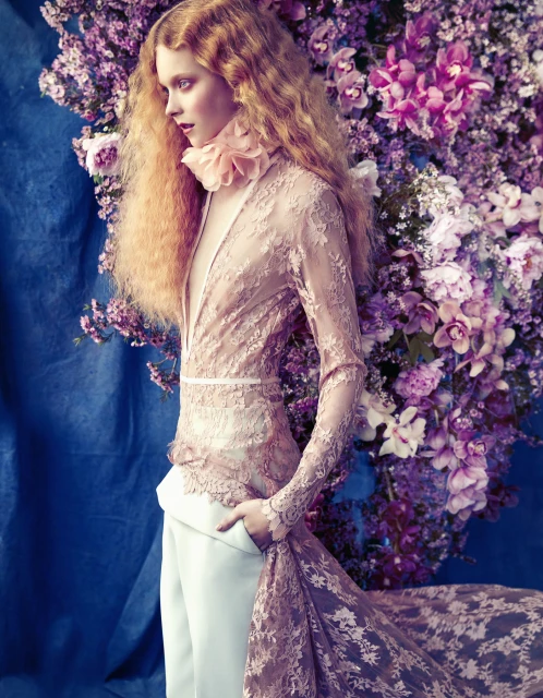 Adult woman in a lace pink long-sleeve dress, light pants, and floral collar poses in front of a blue backdrop and pink and purple flowers.