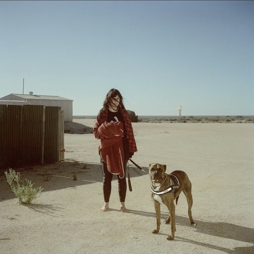 A young woman stands with her brindle dog in a sparse, sun-drenched outdoor setting near a corrugated metal structure on a clear, bright day.