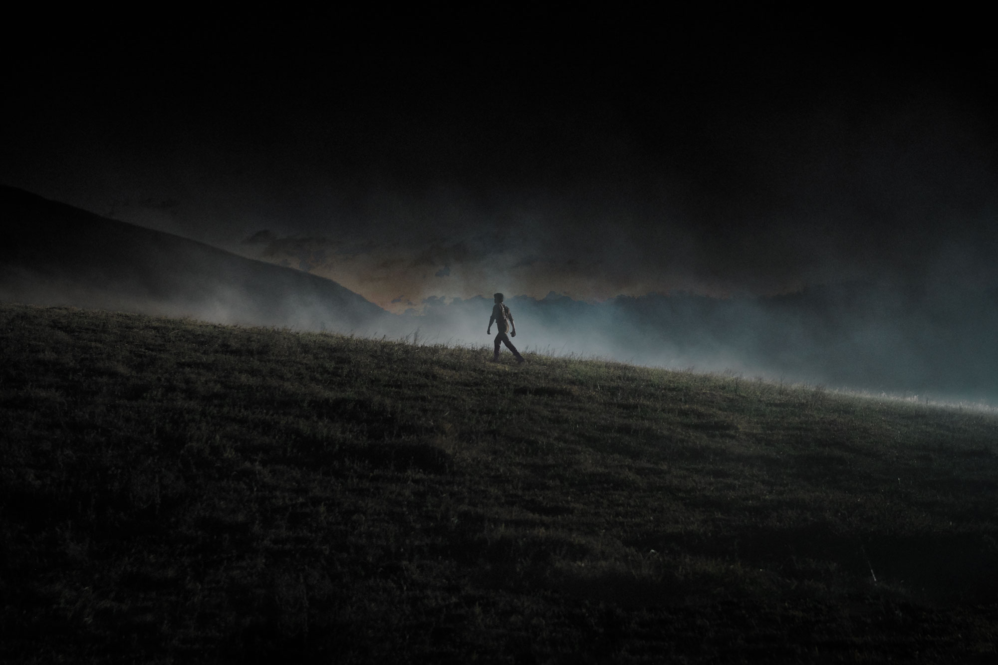 Silhouette of a person walking on a grassy hill under a dark, cloudy sky, creating a moody and dramatic atmosphere.