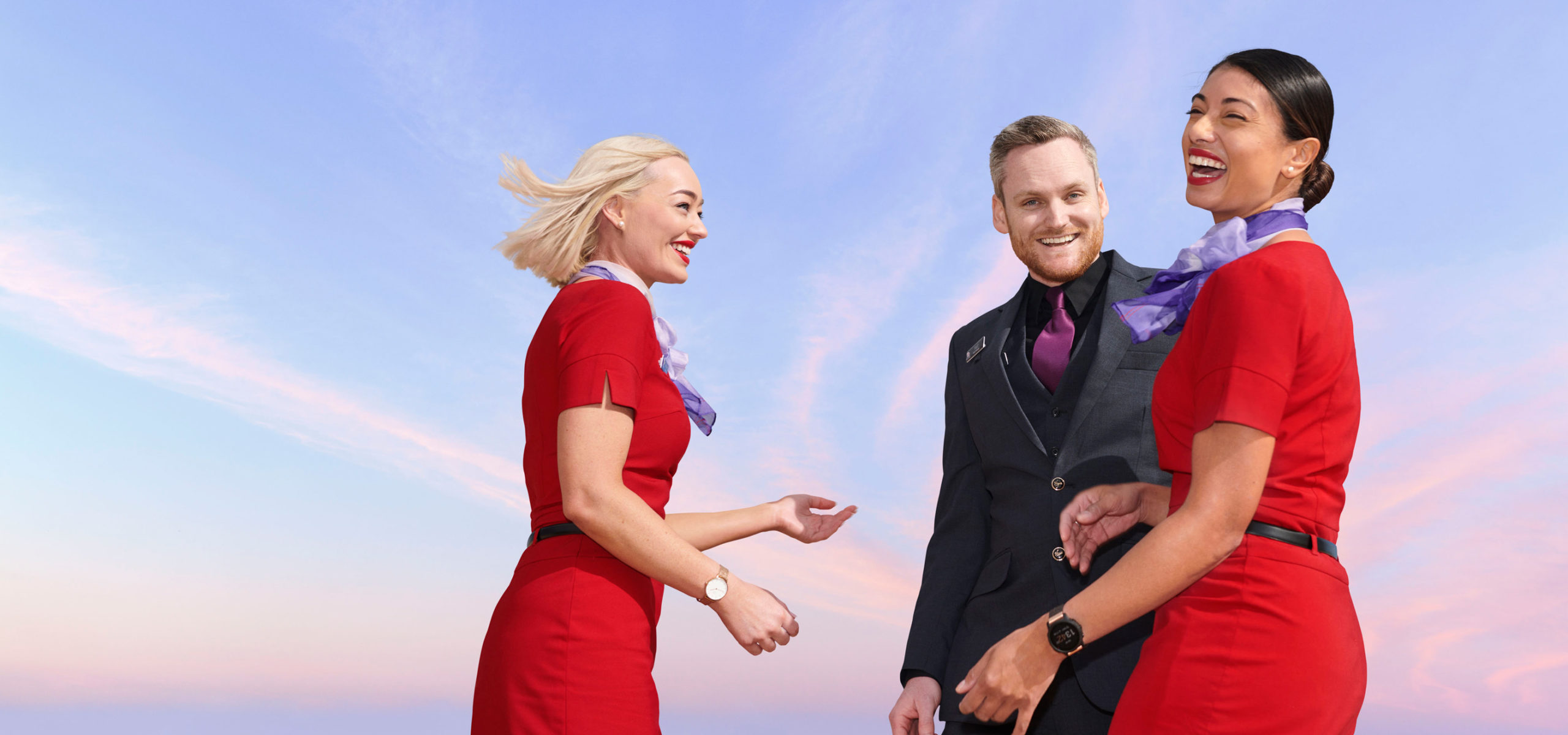 Two smiling women and one man standing outdoors against a bright blue and pink sky, wearing uniforms. Flight attendants, employees, staff.