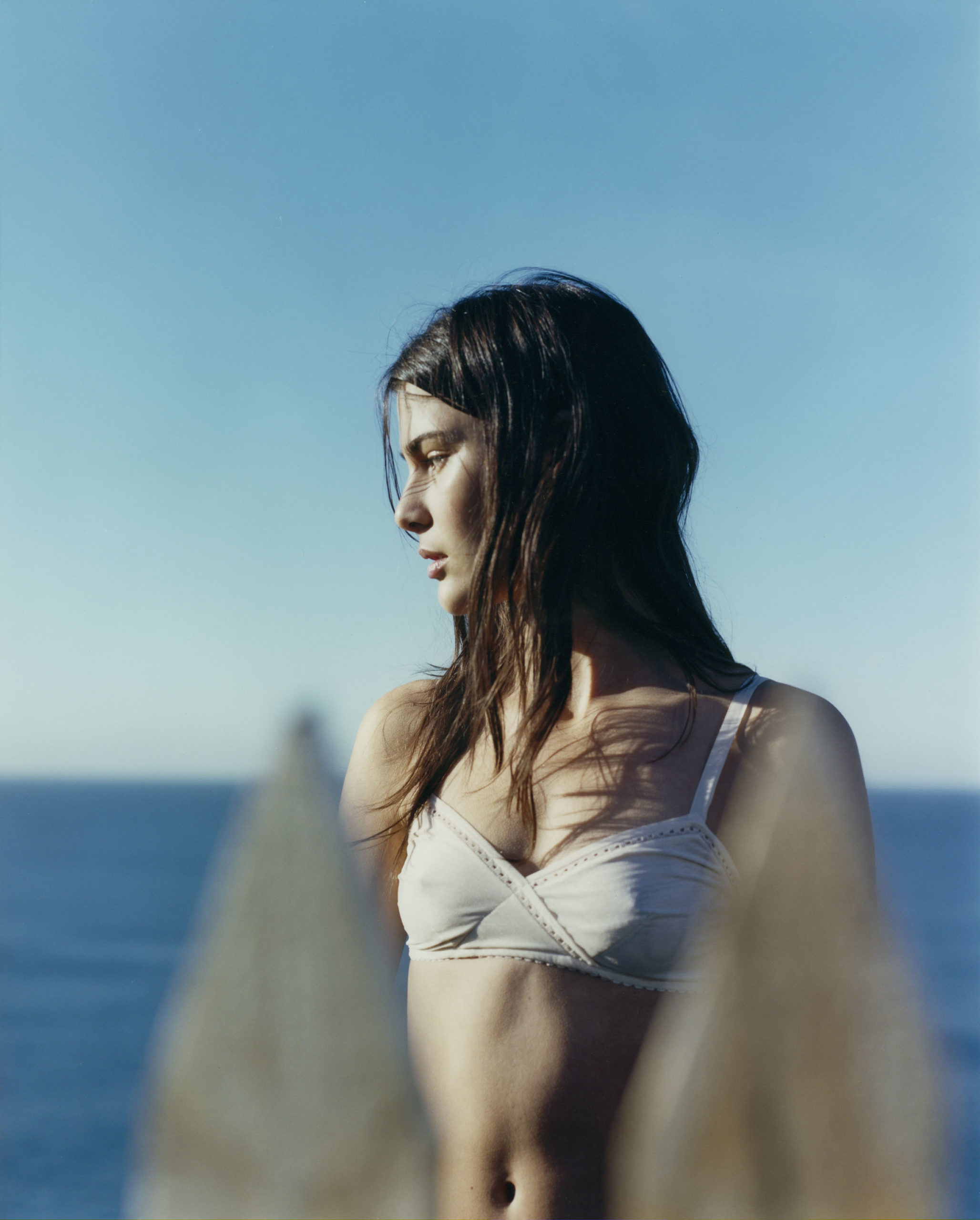 Brunette woman in a bra top poses outdoors with an ocean and blue sky backdrop, capturing a serene and natural aesthetic.