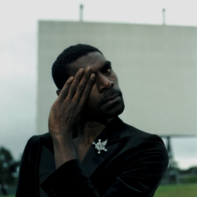 Close-up of an adult male wearing a black jacket with a brooch covering part of his face with his hand, against a blurred outdoor background.