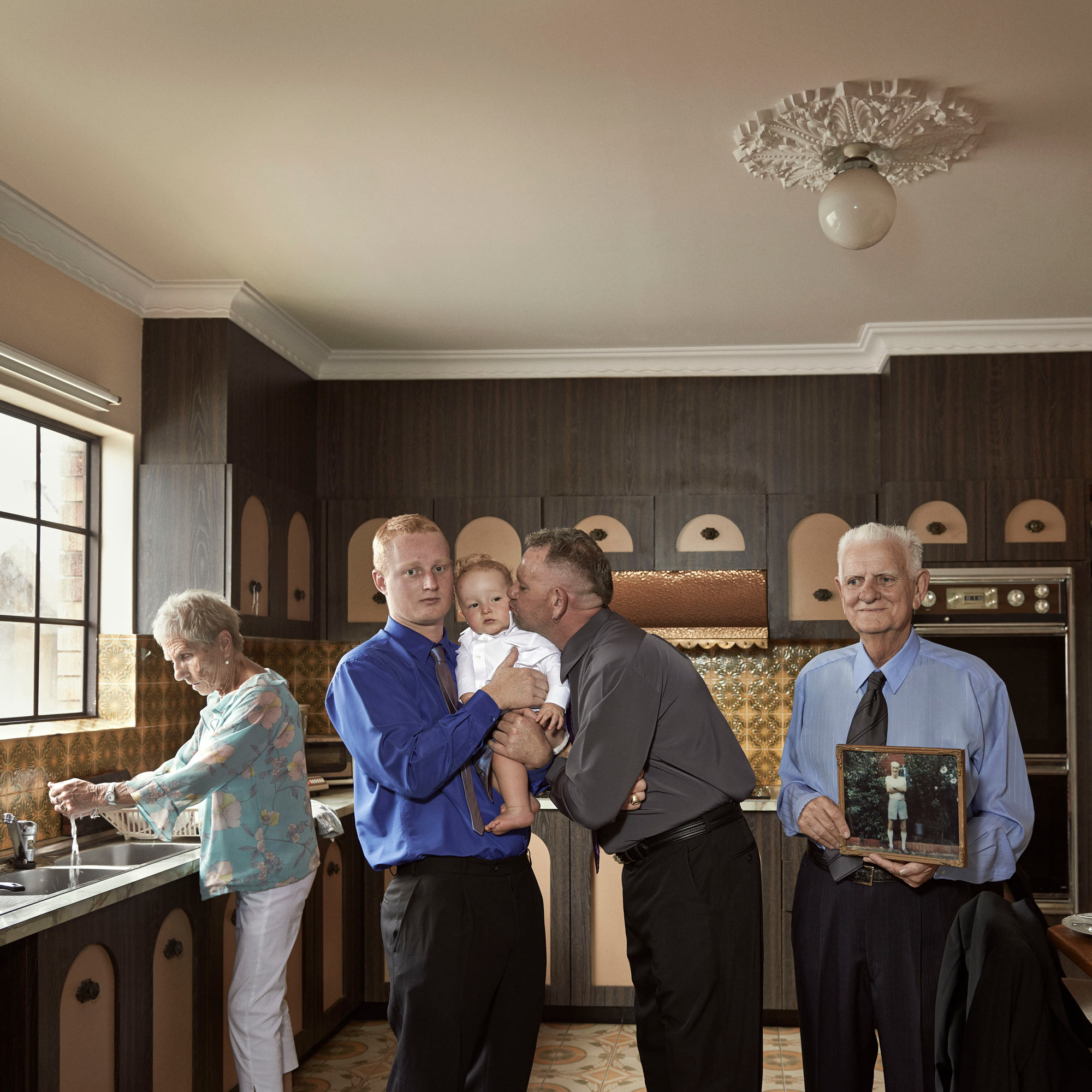 Four adults and a baby in a kitchen, family portrait with one man holding a framed photo, grandma at the sink washing dishes.