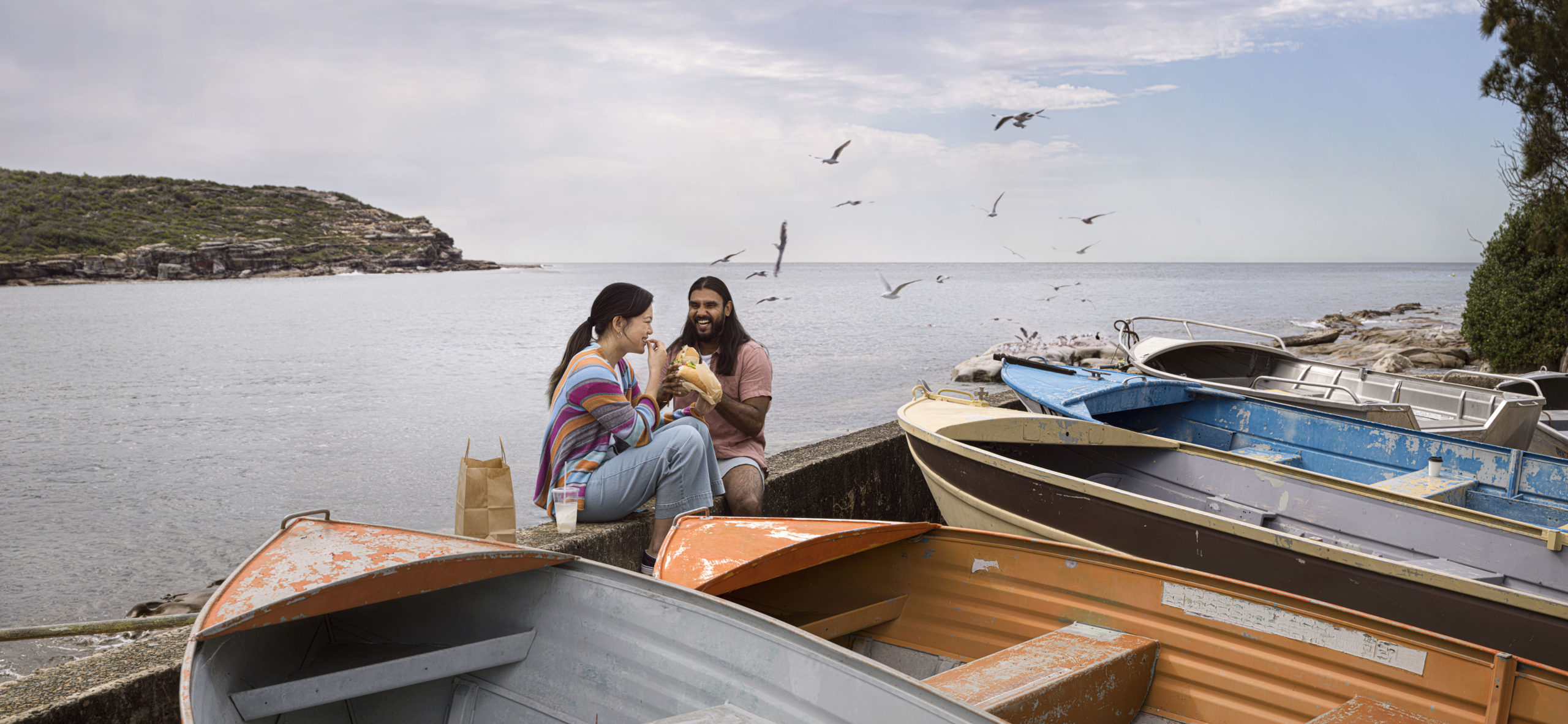 Two people sit by the water eating sandwiches near boats and seabirds under an overcast sky, enjoying a peaceful outdoor meal together.