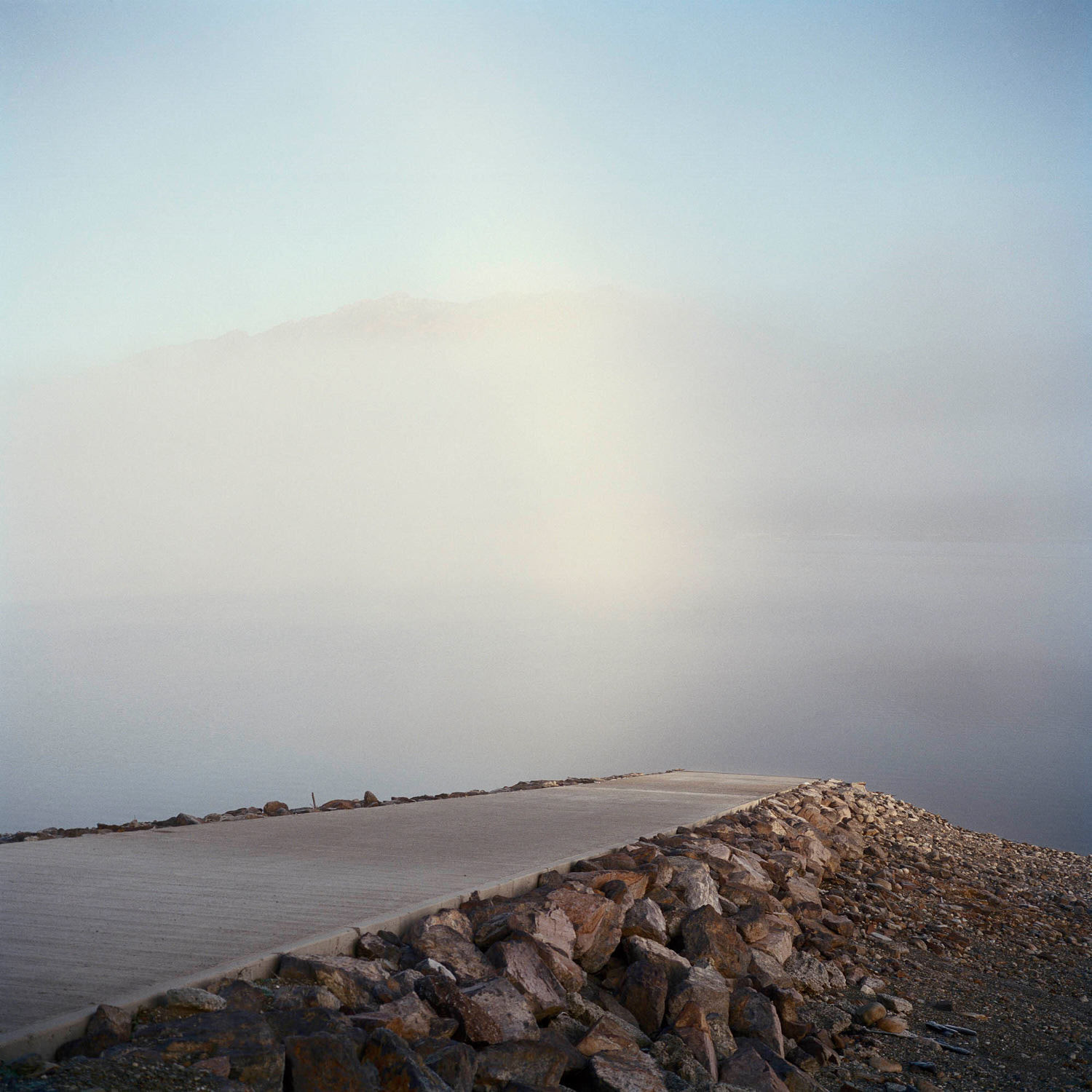 Concrete path along a rocky shoreline disappearing into heavy fog near a body of water under a light blue sky.