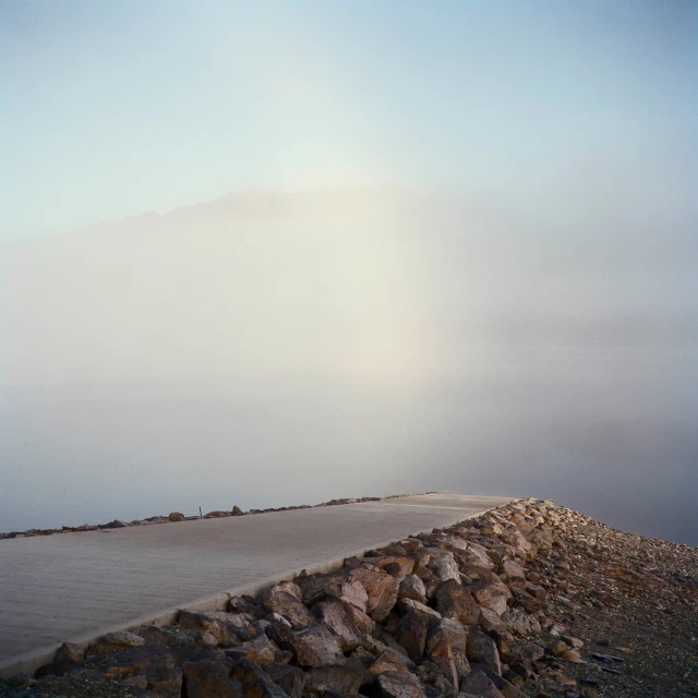 Concrete path along a rocky shoreline disappearing into heavy fog near a body of water under a light blue sky.