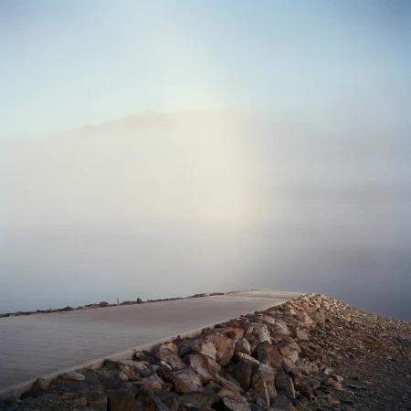 Concrete path along a rocky shoreline disappearing into heavy fog near a body of water under a light blue sky.