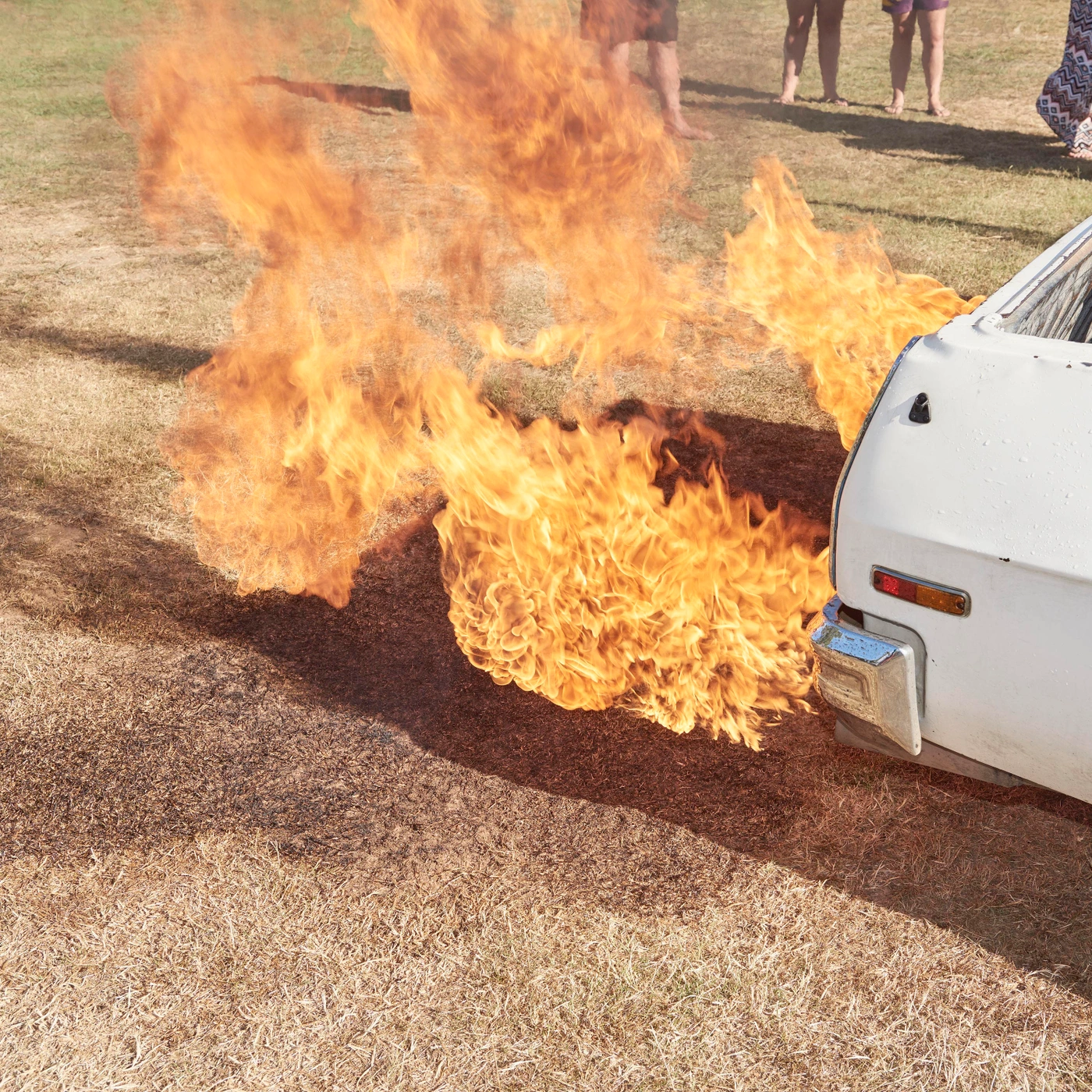 High-angle view of intense bright orange flames erupting near the rear bumper of a white vintage car on dry grass, photographed in Sydney by Ingvar Kenne.