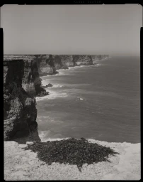 Monochrome film photograph of rugged coastal cliffs meeting the ocean, shot from an elevated viewpoint by Ingvar Kenne.