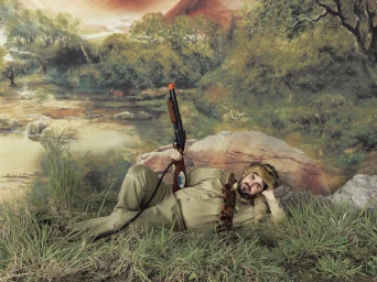 Man wearing a fur hat and khaki shirt reclining in artificial grass holding a toy shotgun against a painted landscape backdrop.