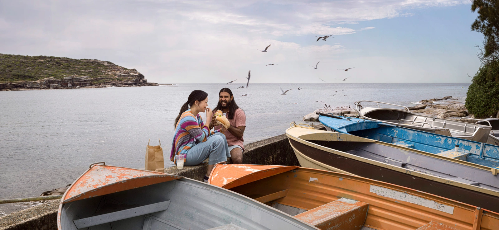Man and woman eating sandwiches on a concrete pier next to small boats with seagulls flying overhead by water.