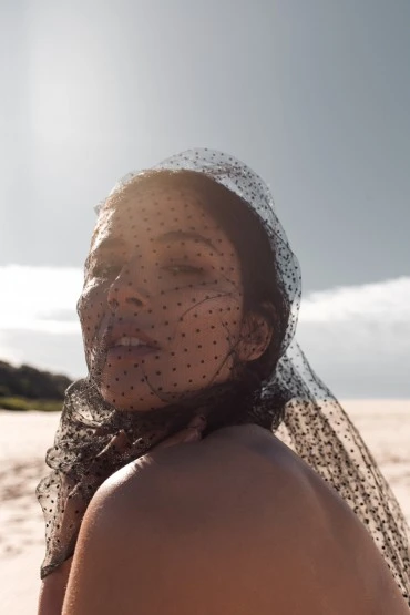 Woman wearing a black dotted veil stands on a sandy beach against a blue sky, shot with natural light for a beauty campaign.