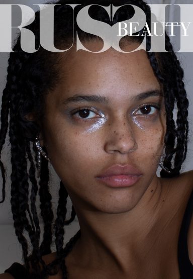 Close-up of an adult female beauty shot with a neutral facial expression, freckles, and a glossy cosmetic application on her face.