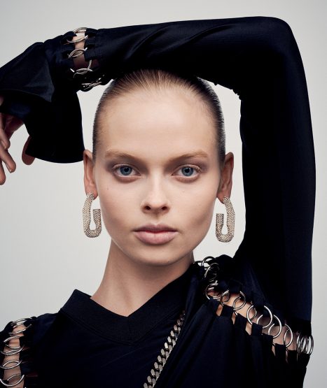 Close-up studio portrait of a female fashion model with geometric earrings wearing a black top with cut-out details, set against a grey backdrop.
