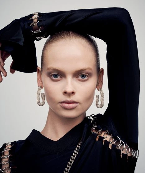 Close-up studio portrait of a female fashion model with geometric earrings wearing a black top with cut-out details, set against a grey backdrop.