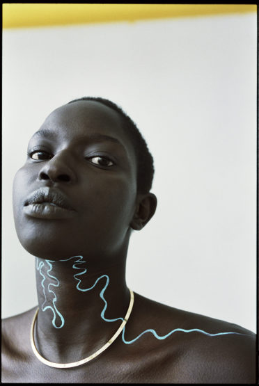 Close-up portrait of a dark-skinned adult female with blue abstract makeup, bare shoulders, and a gold necklace against a white background.
