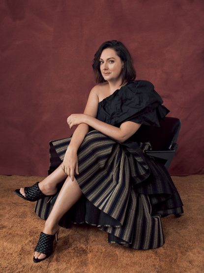 Elegant adult woman in a black one-shoulder top and striped skirt sits in a modern chair against a studio backdrop, showcasing fashion and style.