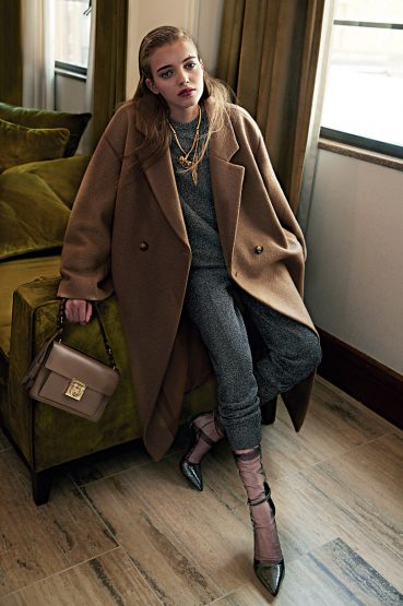 A young adult woman poses indoors wearing a brown overcoat, gray sweater and pants, and black heels, holding a taupe purse.