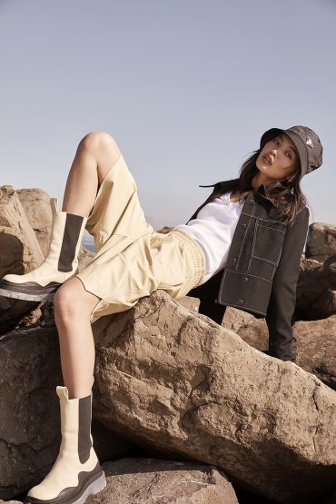 A woman in a bucket hat poses on rocks wearing a white tank top, black jacket, beige shorts, and off-white boots against a blue sky.