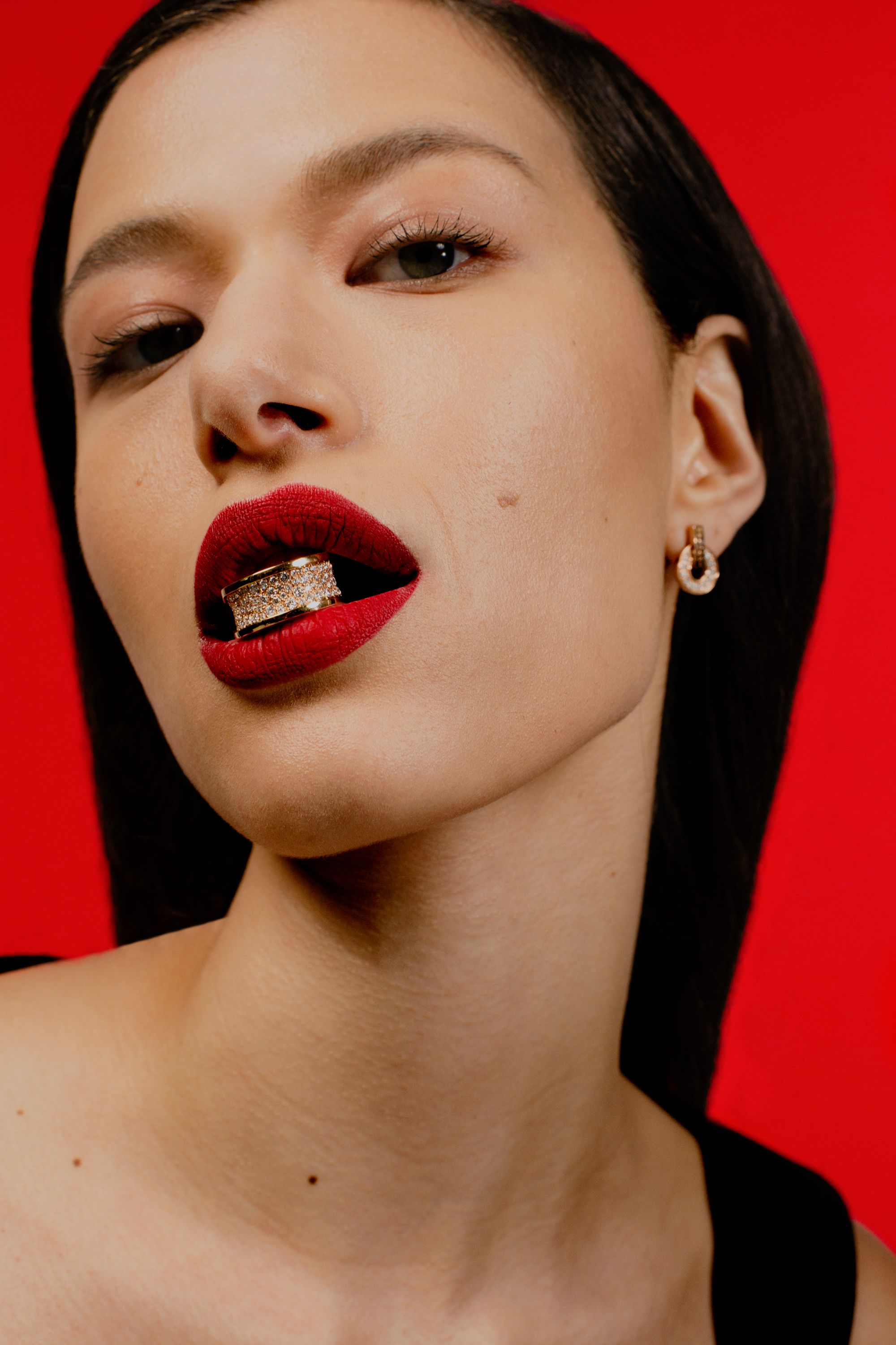 Close-up of a young, light-skinned female model with dark hair wearing red lipstick and holding a jeweled grill in her mouth.