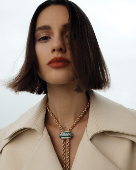 Close-up shot of a young adult woman with short brown hair wearing an off-white trench coat and a decorative gold pendant necklace.