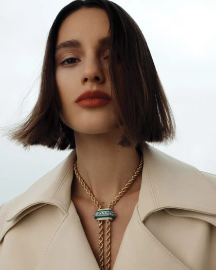 Close-up shot of a young adult woman with short brown hair wearing an off-white trench coat and a decorative gold pendant necklace.