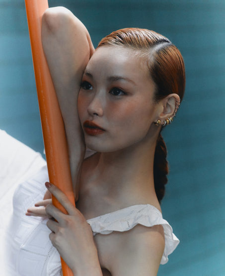 Close-up portrait of an Asian woman with copper-red hair, leaning against an orange bar, with a blurred blue background.