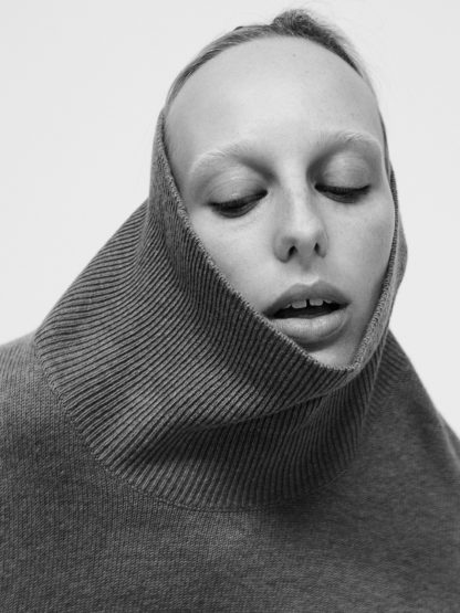Close-up portrait of a woman wearing a grey knit turtleneck pulled up over her chin, with makeup by Filomena Natoli.
