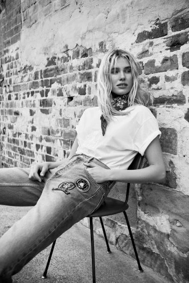 Black and white shot of a stylish adult woman with wavy blonde hair sitting on a metal chair in front of a brick wall.