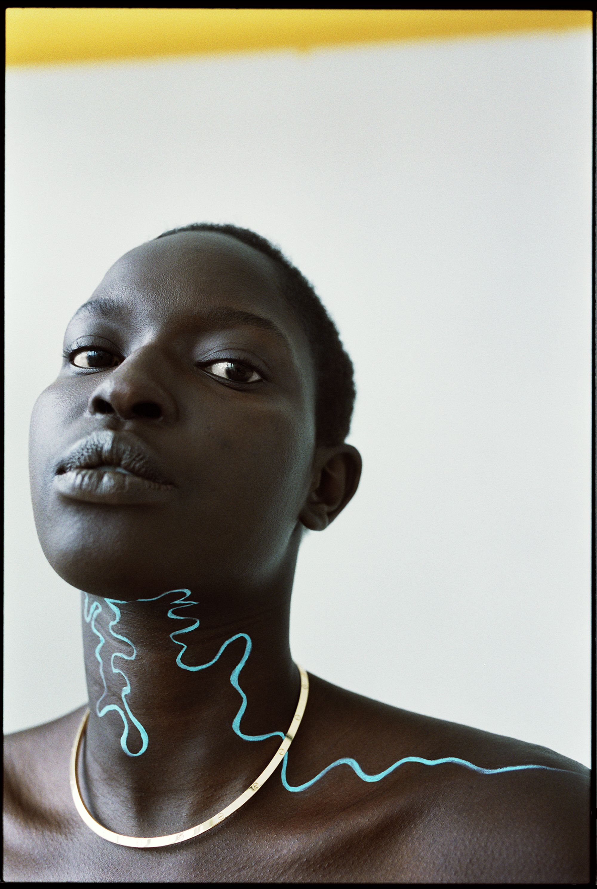 Close-up portrait of a Black female model with turquoise paint on her neck and a gold necklace against a white background.