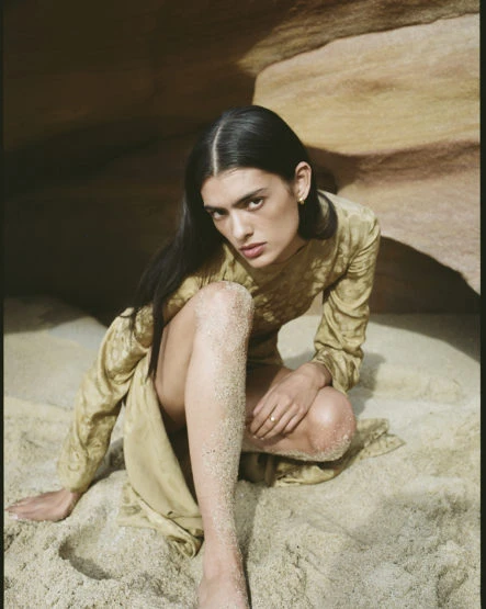 Woman in long-sleeve beige dress sits on sand with tan rock formation in the background, shot in a medium, eye-level camera angle.