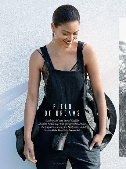 Model with slicked back hair wearing black overalls over a lace top against a white textured wall