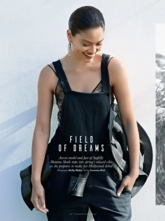 Model with slicked back hair wearing black overalls over a lace top against a white textured wall