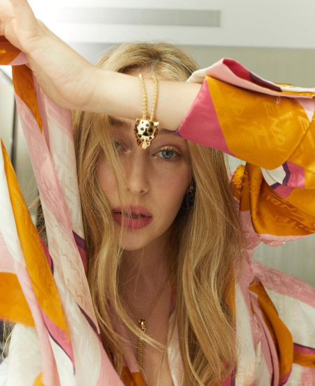 Close-up of a woman with wavy blonde hair wearing a gold necklace with a jaguar pendant and a pink, white and orange patterned shirt.