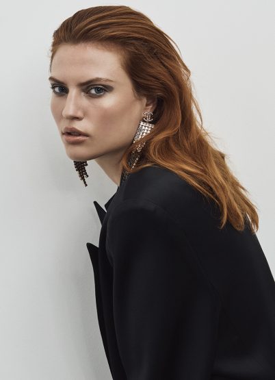 Portrait of a red-haired woman in a black jacket wearing crystal drop earrings against a neutral background.