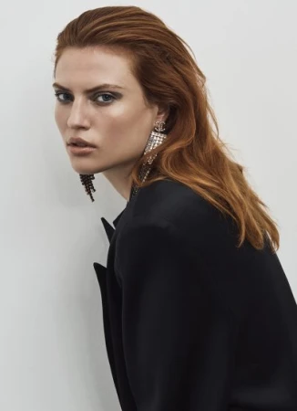 Portrait of a red-haired woman in a black jacket wearing crystal drop earrings against a neutral background.