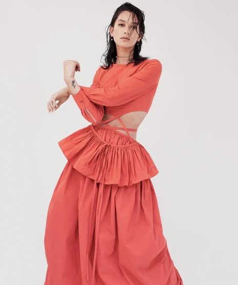 Fashionable young woman poses in the photo studio wearing a coral-colored tiered dress with a bare midriff against a neutral backdrop.