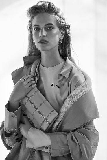 Monochrome portrait of a young woman wearing a t-shirt, open shirt, and jacket, holding a clutch purse in a brightly lit indoor setting.