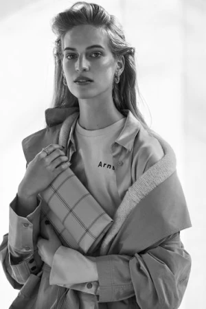 Monochrome portrait of a young woman wearing a t-shirt, open shirt, and jacket, holding a clutch purse in a brightly lit indoor setting.