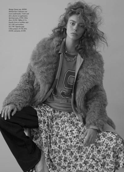 Fashion editorial portrait: Woman in oversized coat and floral skirt sits against a grey background in a studio, shot in black and white.