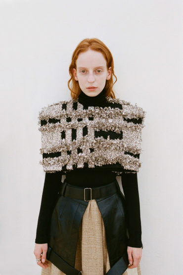 Fashion model in a black turtleneck, embellished cape, black belt, and beige skirt, posing in front of a white background.
