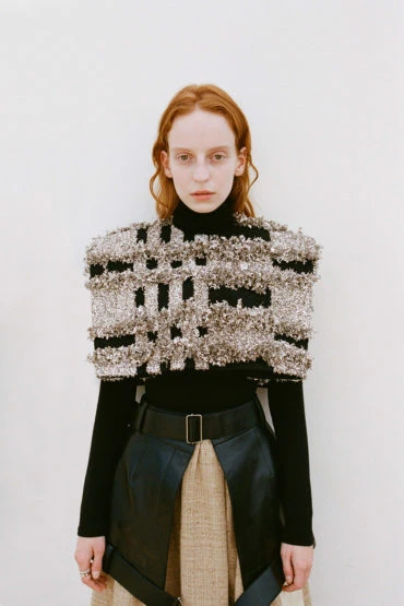 Fashion model in a black turtleneck, embellished cape, black belt, and beige skirt, posing in front of a white background.