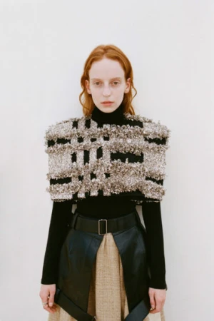 Fashion model in a black turtleneck, embellished cape, black belt, and beige skirt, posing in front of a white background.