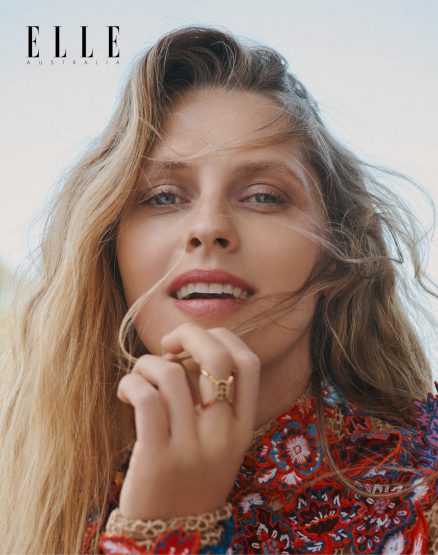 Close-up portrait of a blonde woman with hand near face wearing a colorful patterned top in an outdoor setting for Elle Australia.