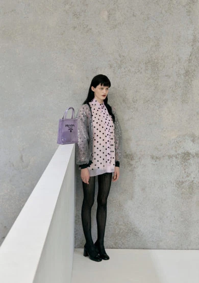 Full shot of a young adult female fashion model in a polka dot dress, tights, and heels next to a lavender Prada handbag.