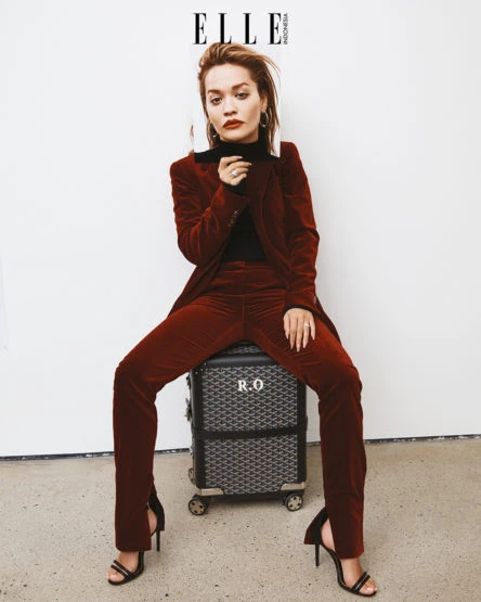 Woman in a red velvet suit sits on a patterned suitcase with black heels in a studio setting for an Elle Indonesia fashion editorial.