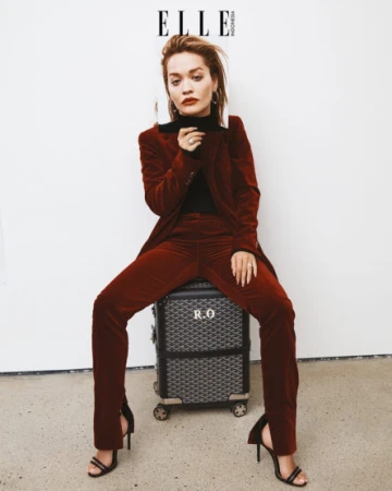 Woman in a red velvet suit sits on a patterned suitcase with black heels in a studio setting for an Elle Indonesia fashion editorial.